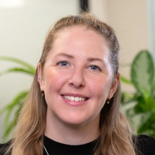 Portrait of a smiling woman with blond hair in an office, plants in the background.