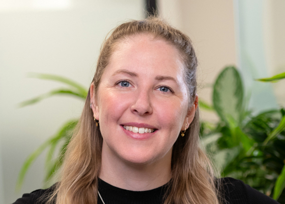 Portrait of a smiling woman with blond hair in an office, plants in the background.