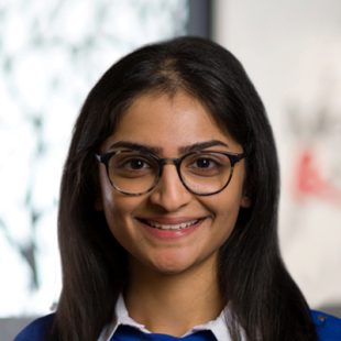 Portrait of a young woman with dark hair and glasses smiling at the camera in a bright indoor setting.