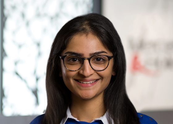 Portrait of a young woman with dark hair and glasses smiling at the camera in a bright indoor setting.