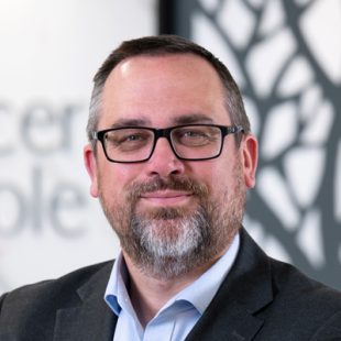 Portrait of a middle-aged man with glasses and a beard, wearing a dark blazer and light blue shirt, in a modern office setting.