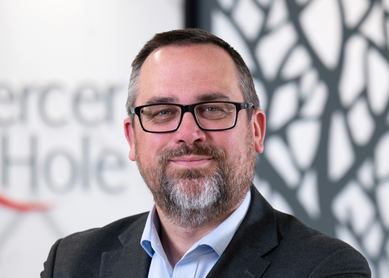 Portrait of a middle-aged man with glasses and a beard, wearing a dark blazer and light blue shirt, in a modern office setting.