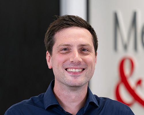 Head-and-shoulders portrait of a man in a blue shirt smiling at the camera in an office setting.