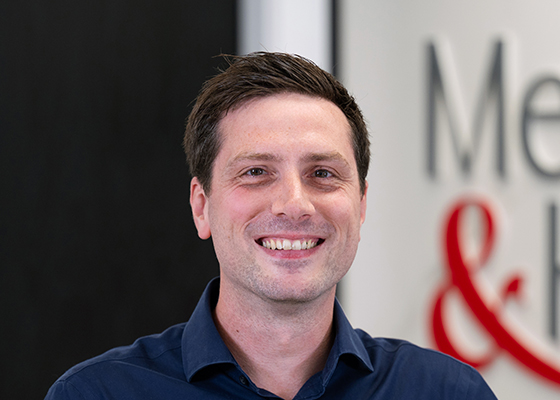 Head-and-shoulders portrait of a man in a blue shirt smiling at the camera in an office setting.